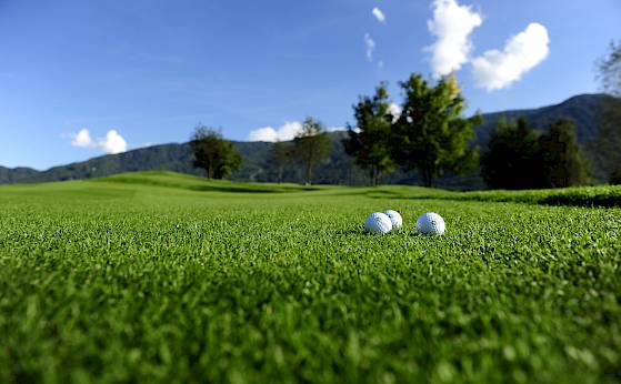 Grüner Golfplatz mit zwei Golfbällen, umgeben von Bäumen und Bergen unter blauem Himmel.