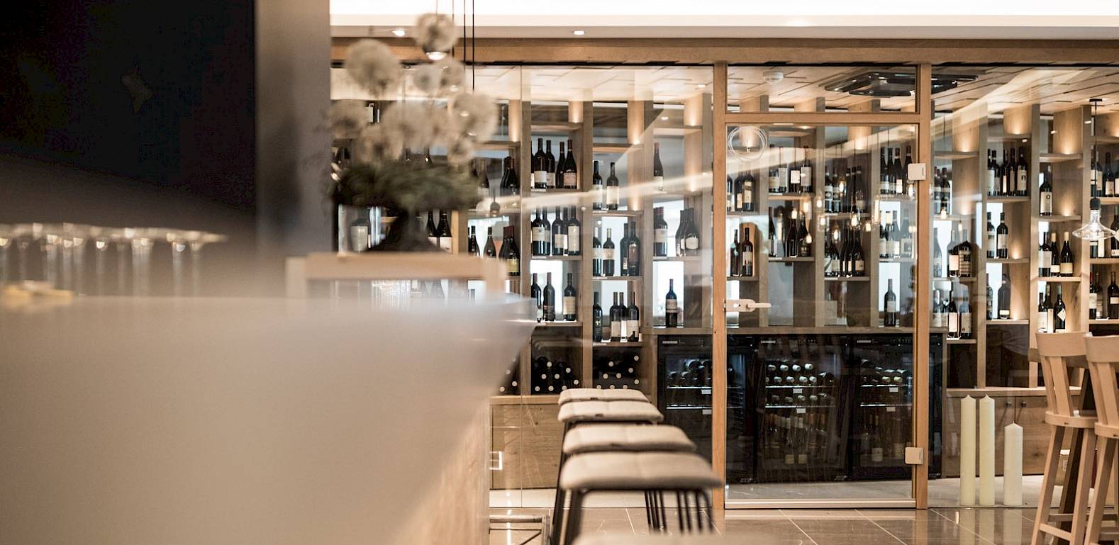 Enoteca moderna con scaffali in legno pieni di bottiglie, bancone e sgabelli, illuminazione suggestiva.