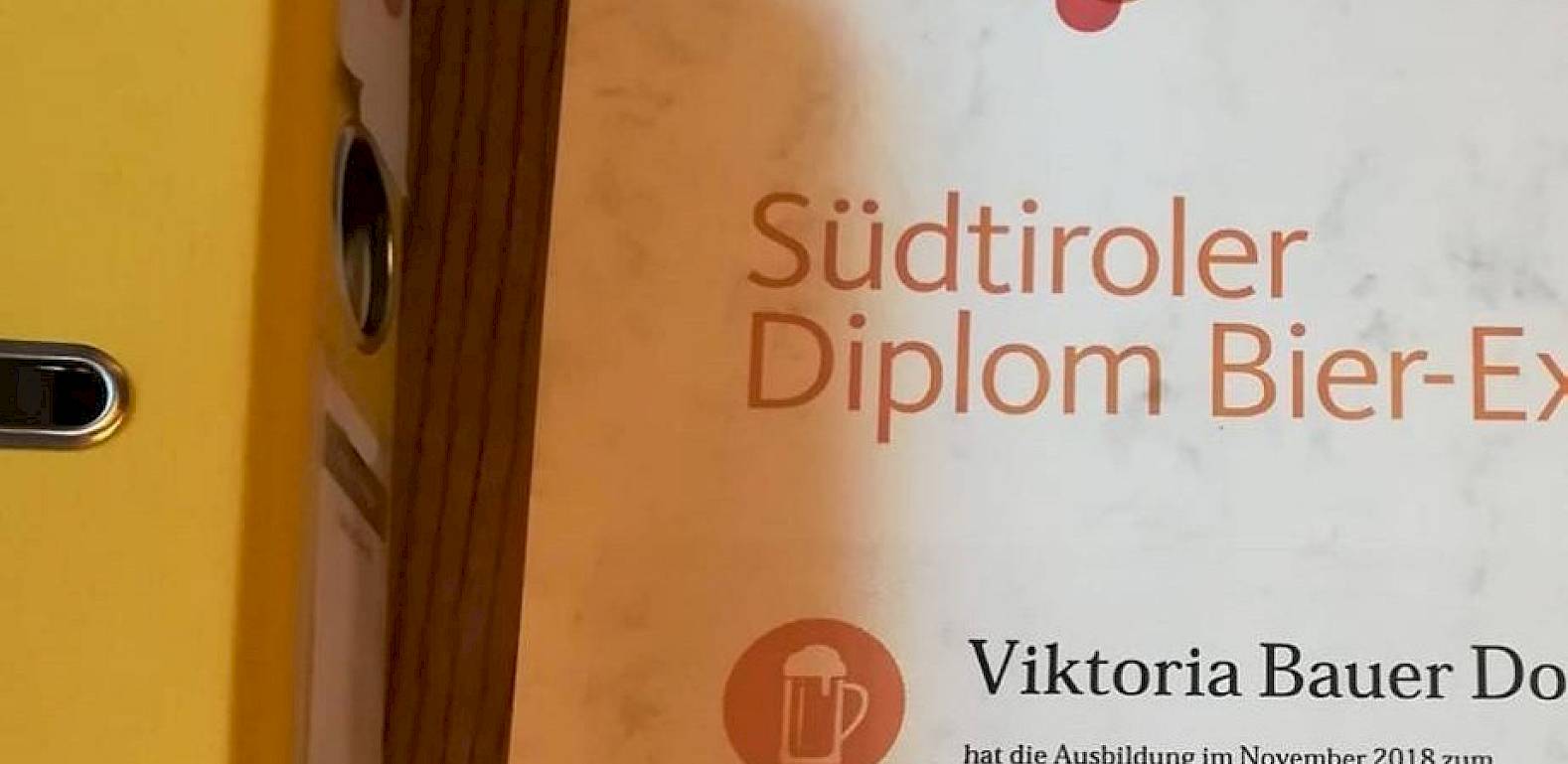 Un certificato dell'Accademia della Birra dell'Alto Adige per esperti di birra accanto a una cartella gialla.