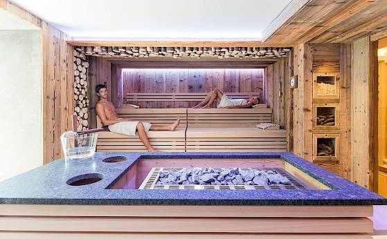 Eine Holzsauna mit zwei entspannenden Personen auf Bänken, ein Wasserkrug und Saunaofen im Vordergrund.