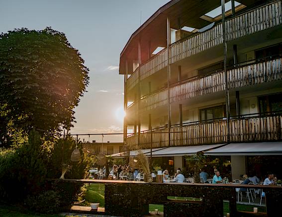 Moderne Holzgebäude, Abendsonne, Menschen speisen draußen im Garten, entspannte Atmosphäre.