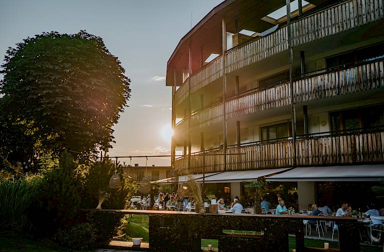 Moderne Holzgebäude, Abendsonne, Menschen speisen draußen im Garten, entspannte Atmosphäre.