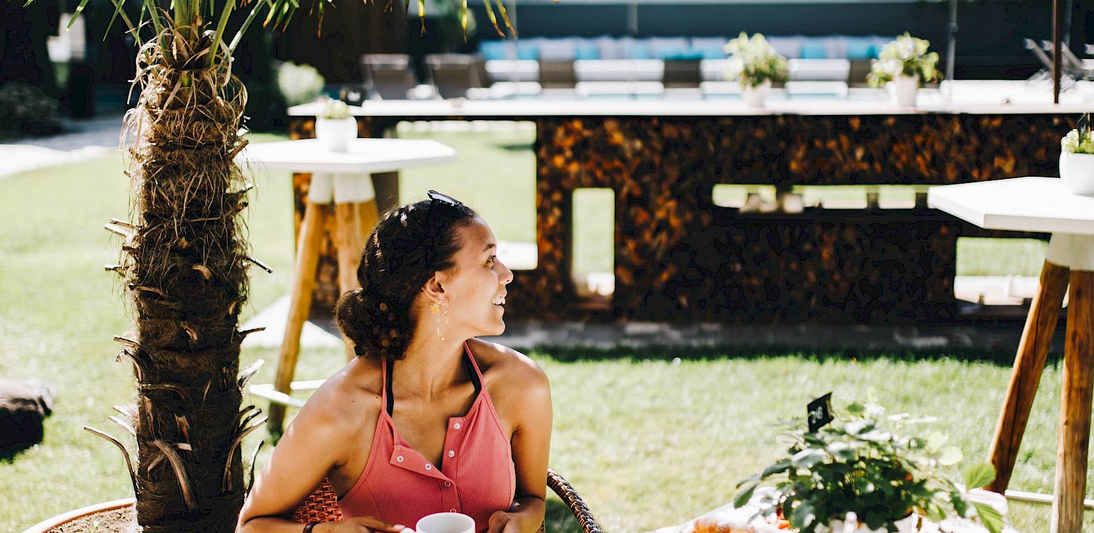 Woman in red dress enjoys breakfast outdoors by a palm, surrounded by green landscape.