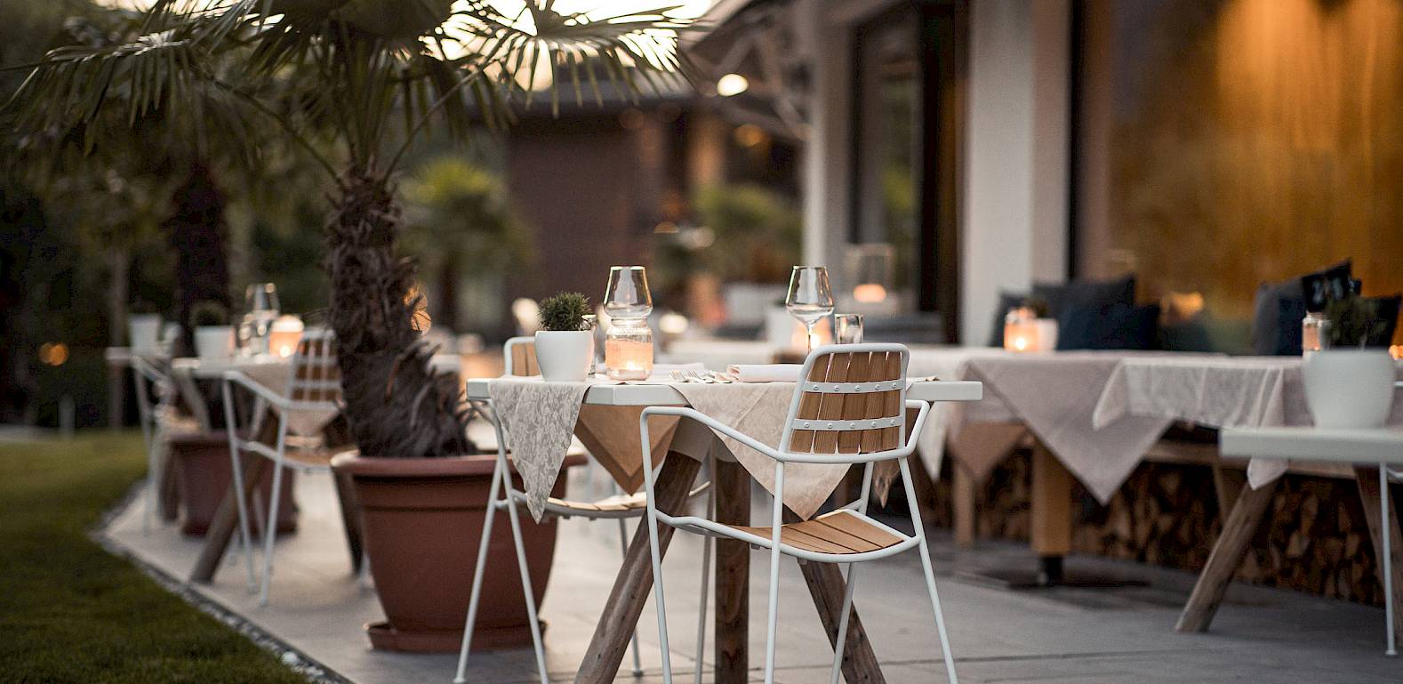 Stylish restaurant outdoor area with tables, chairs, and potted palm trees at sunset.