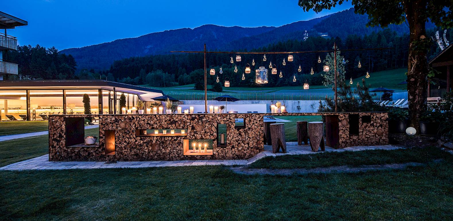 Illuminated outdoor bar made of logs at dusk, surrounded by picturesque mountain scenery.
