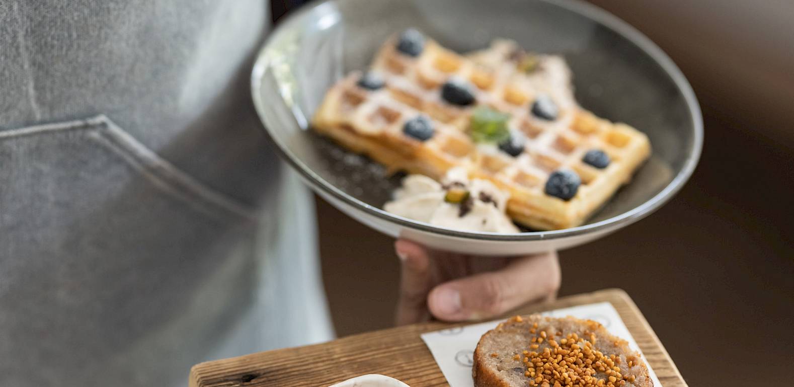 Person in Schürze hält Teller mit Waffeln und eine Holzplatte mit Brot und Honigschälchen.