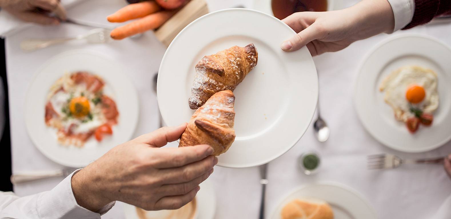 Ein Frühstückstisch mit Croissants, Obst und Rührei. Zwei Personen teilen ein Croissant auf einem Teller.