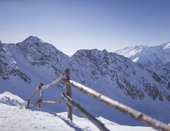 Snowy mountain landscape with wooden fence, blue sky, and sunlit peaks in the background.