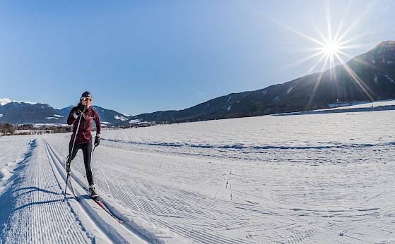 Skifahrer auf gespurter Loipe unter klarem Himmel, Berge im Hintergrund, Sonne strahlt hell.