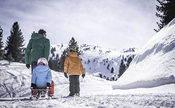 Familie zieht Schlitten im verschneiten Berggebiet, umgeben von Bäumen und strahlend blauem Himmel.