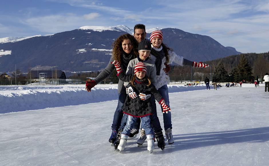 Familie beim Schlittschuhlaufen auf einem zugefrorenen See mit schneebedeckten Bergen im Hintergrund.