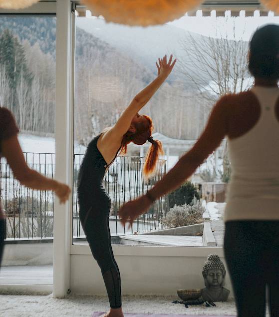Drei Personen machen Yoga in einem hellen Raum mit Blick auf verschneite Hügel. Entspannte Atmosphäre.