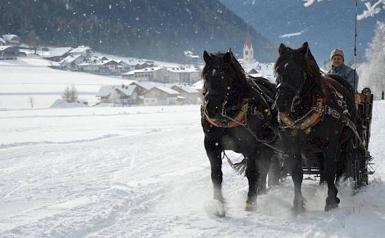 Pferdeschlitten mit zwei schwarzen Pferden im Schnee, vor einem winterlichen Dorf und Kirche im Hintergrund.