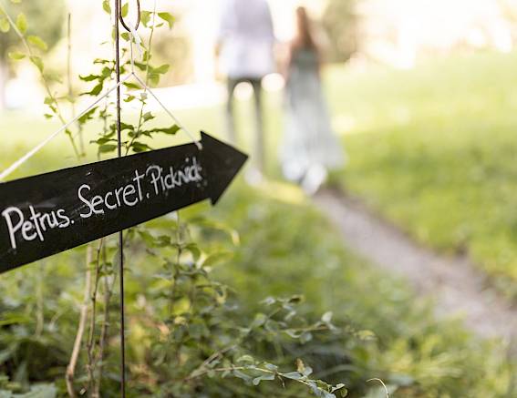 Cartello nero tra l'erba con scritto "Petrus Secret Picknick", una coppia sul sentiero sullo sfondo.