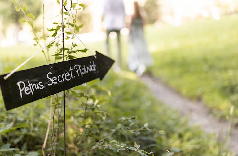 Cartello nero tra l'erba con scritto "Petrus Secret Picknick", una coppia sul sentiero sullo sfondo.