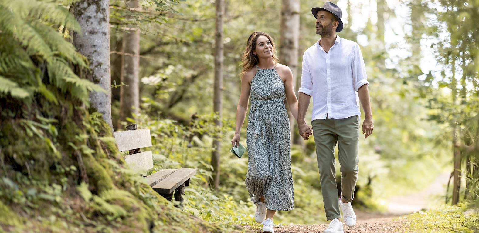A couple walking on a forest path, smiling, surrounded by trees and ferns, on a sunny day.