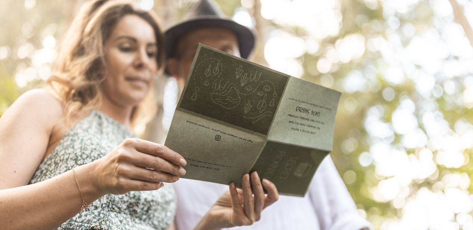 A woman and a man read a brochure in the woods. They stand close together and appear relaxed.