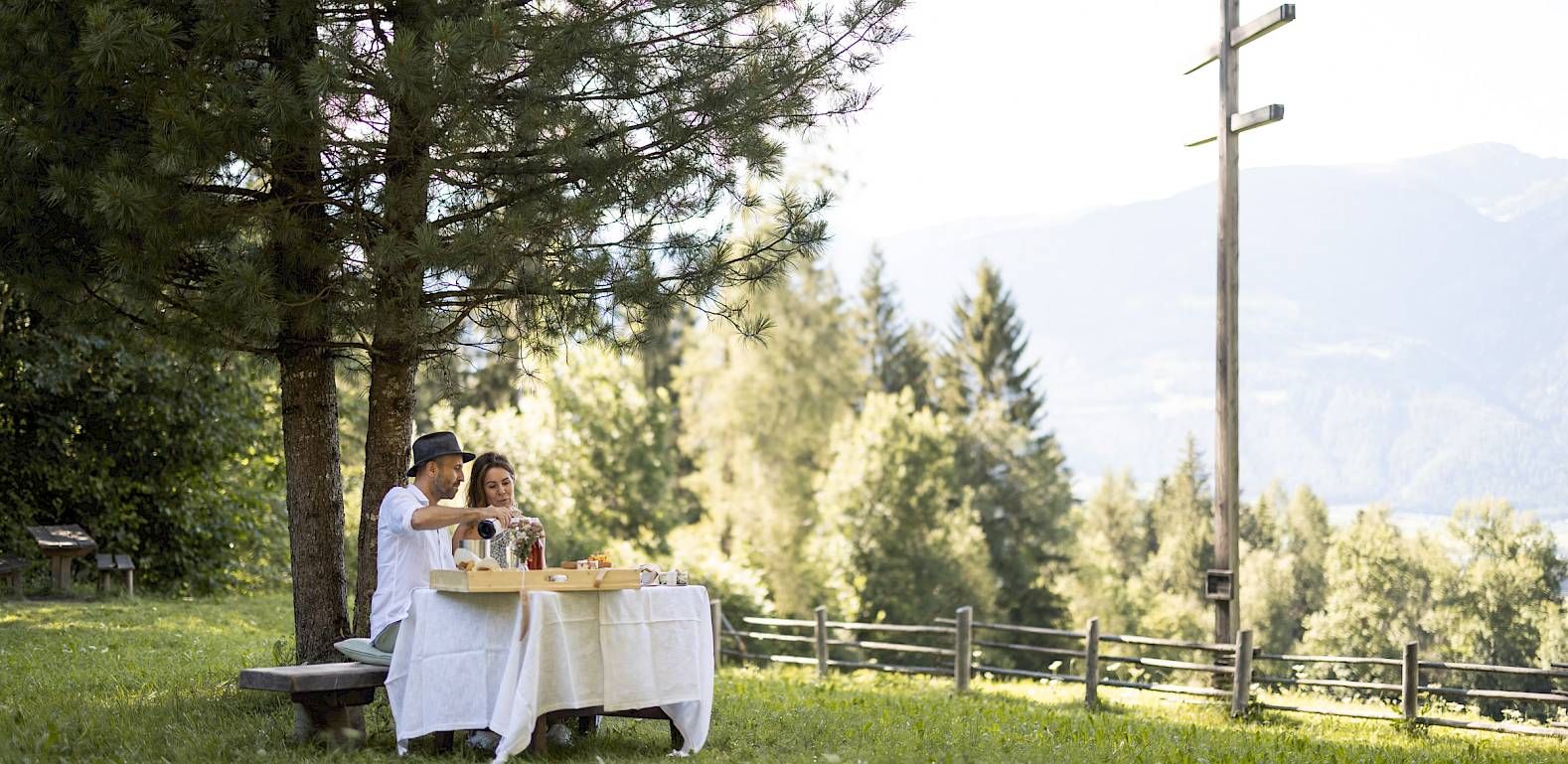 A couple picnics under a tree in a scenic mountain landscape, with pine trees and an open sky.