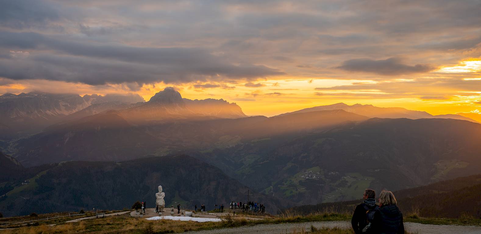 Zwei Menschen bei Sonnenuntergang in Berglandschaft, mit Statue und Wolkenhimmel im Hintergrund.