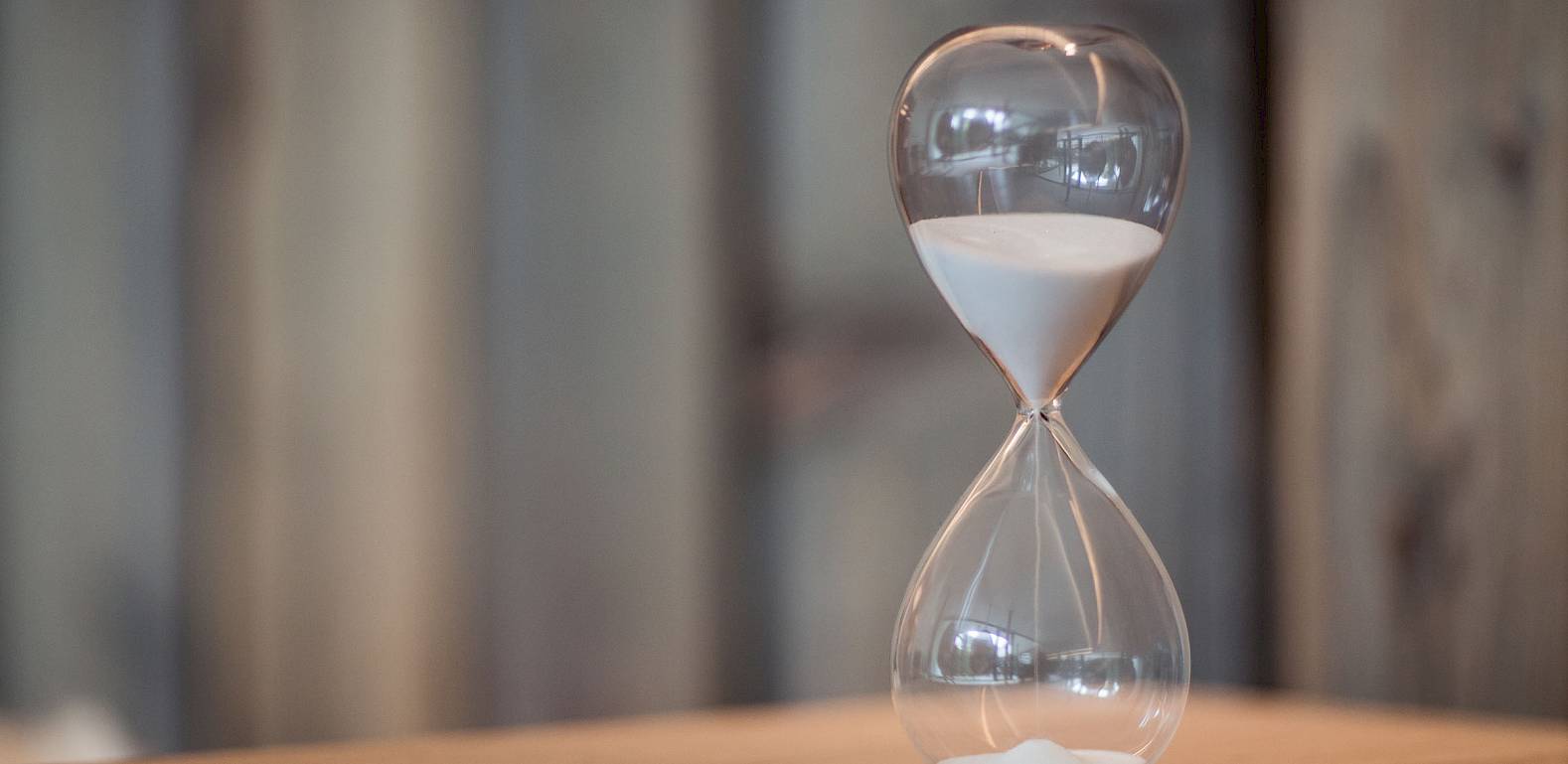 Eine Sanduhr mit feinem, weißem Sand zeigt die fließende Zeit auf einem Holztisch vor hölzernem Hintergrund.