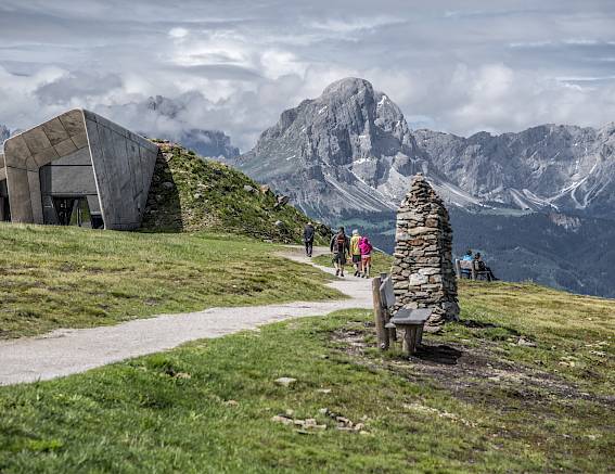 Wanderer auf einem Gebirgspfad mit einer modernen Steinskulptur, umliegenden Bergen und Wolken im Hintergrund.