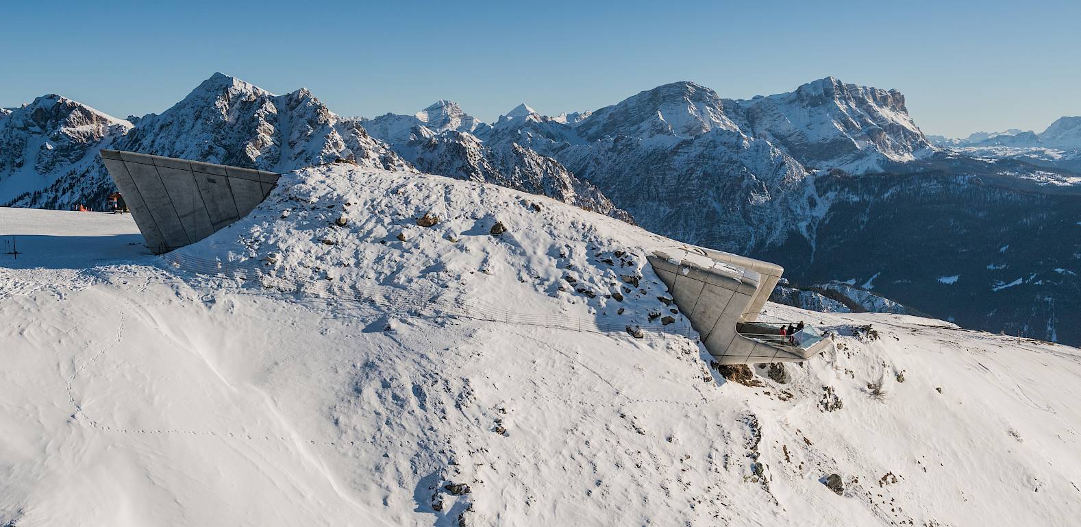 Mountain panorama with snow-covered peaks and modern, angular structures under a blue sky.