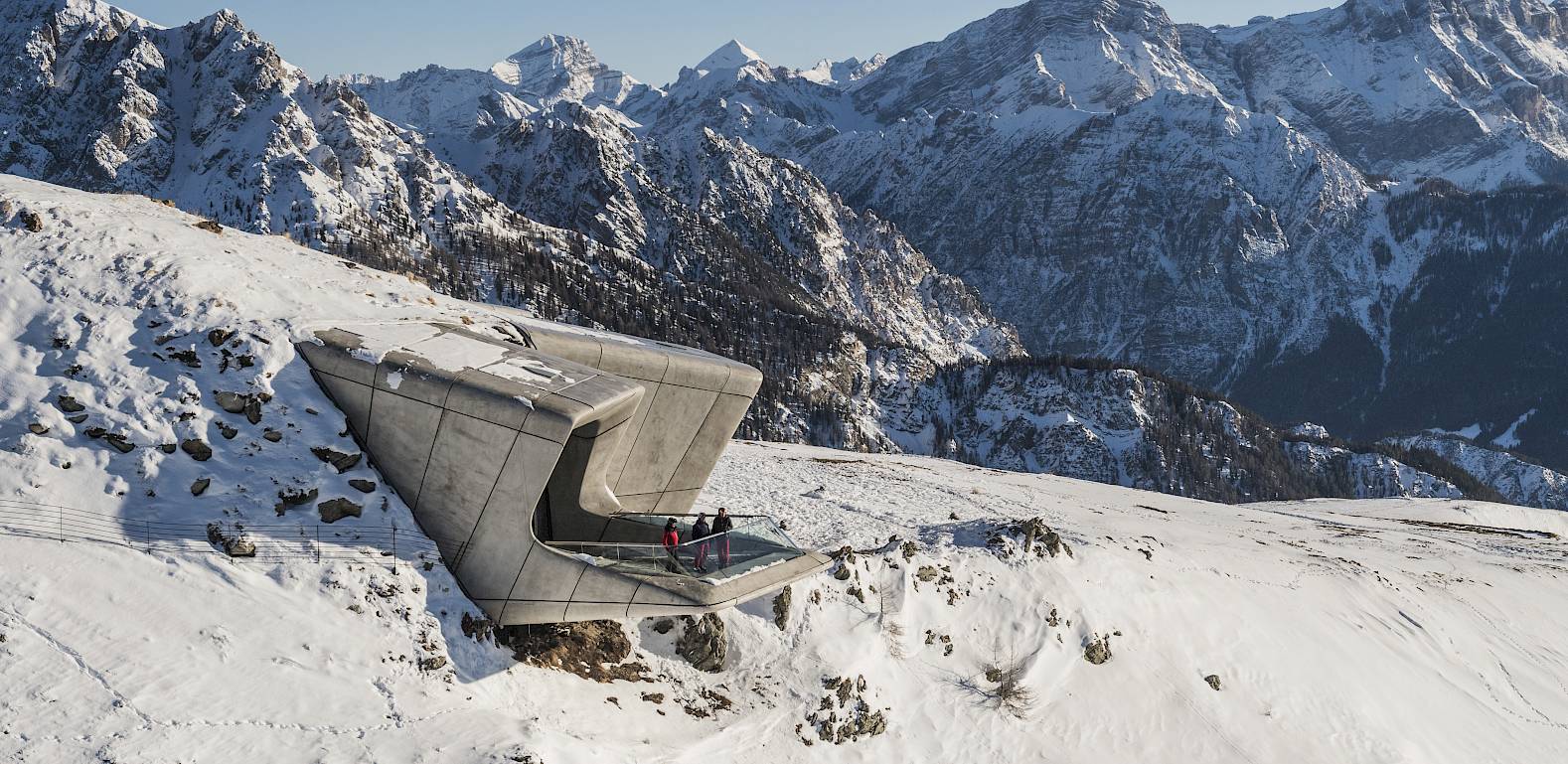 Mountain museum nestled in snowy slope, surrounded by jagged mountain peaks under a clear sky.