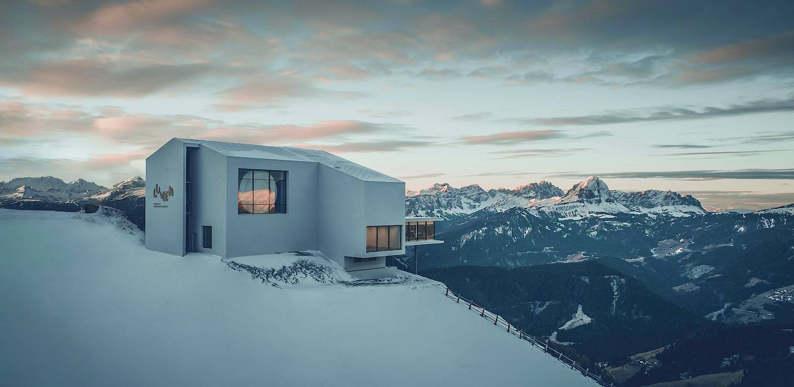 Mountain house on snow-covered peak at sunset, overlooking panoramic alpine landscape.