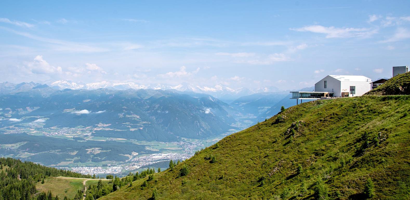 Modern mountain hut on a green slope overlooking a wide valley and snowy Alpine peaks in the distance.