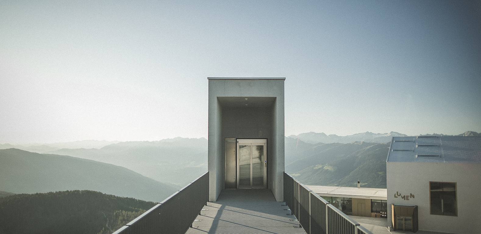 A modern viewing tower with a wide mountain view under a clear sky. Building in the foreground.