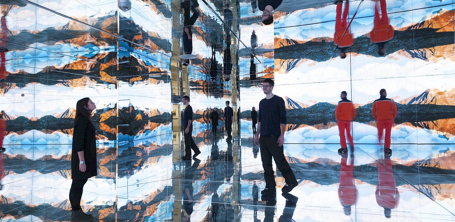A room full of mirrors with mountain panorama, two people in black and red explore a fascinating scene.