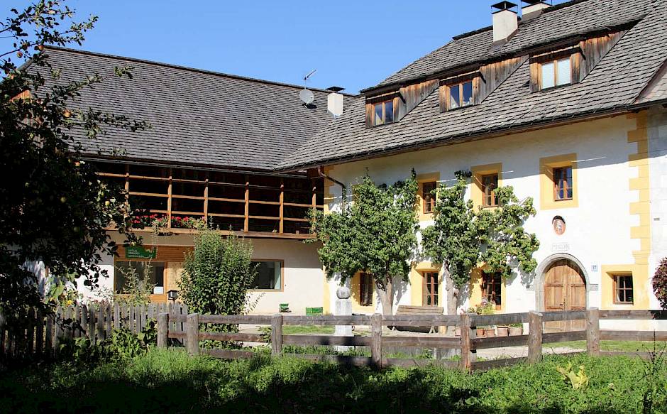 Traditionelles Bauernhaus mit weißer Fassade, gelben Fenstern, Holzbalkon und Gartenzaun im Sonnenschein.