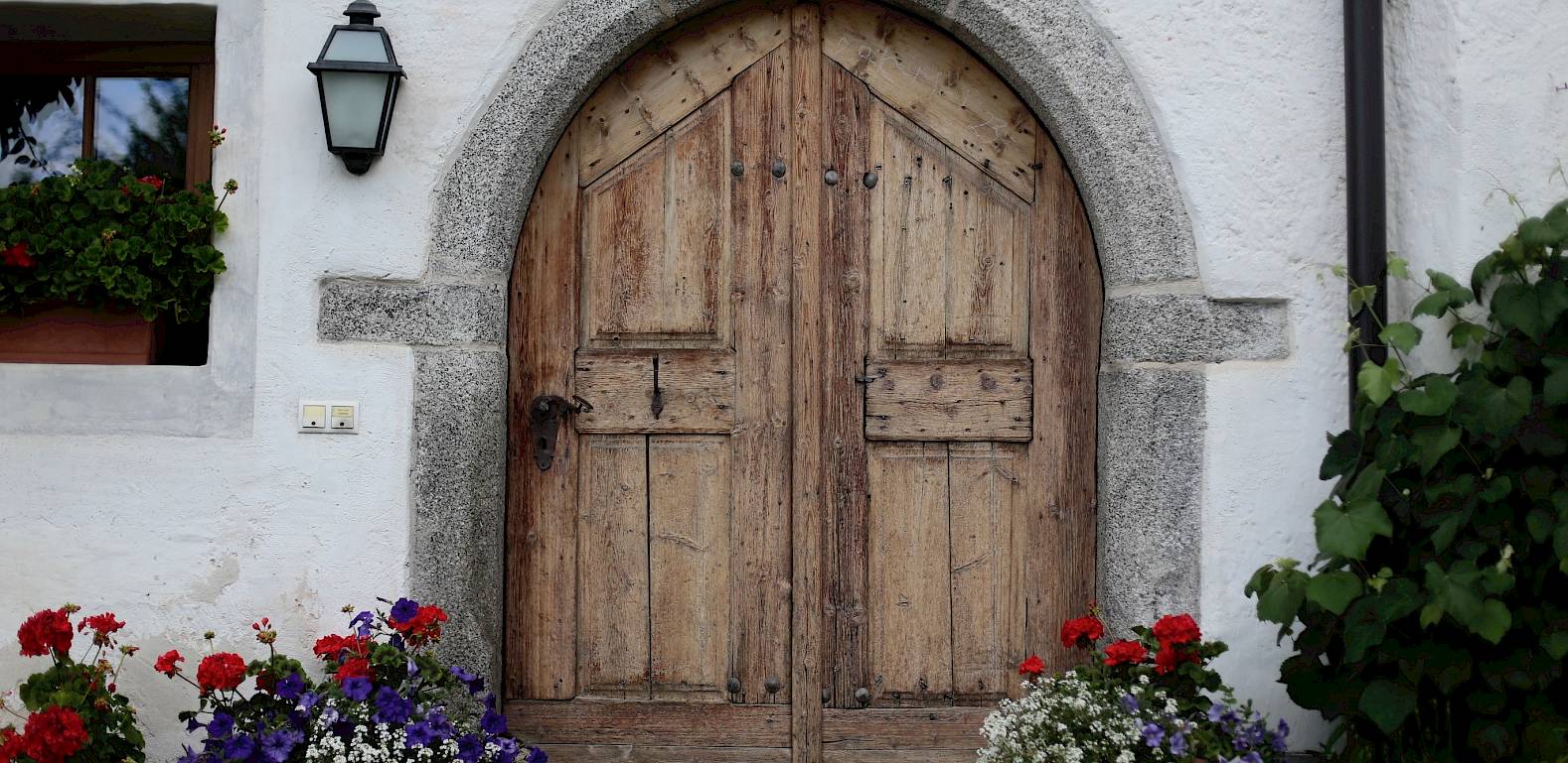 Porta in legno con arco in pietra, circondata da fiori in vaso e una lanterna su facciata bianca.