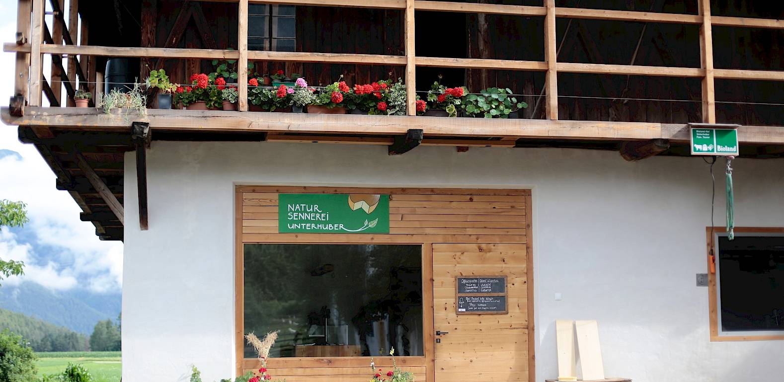 Casa in legno con veranda in legno, vasi di fiori e campi verdi sullo sfondo. Cartello: "Centro Natura e Avventura".