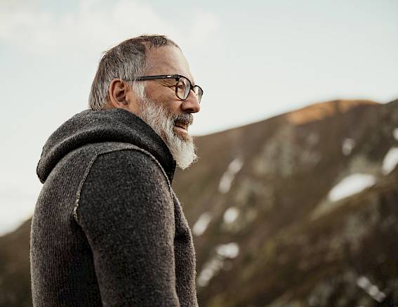 Uomo anziano con occhiali e maglione grigio davanti a paesaggio montano boscoso, visto di lato.