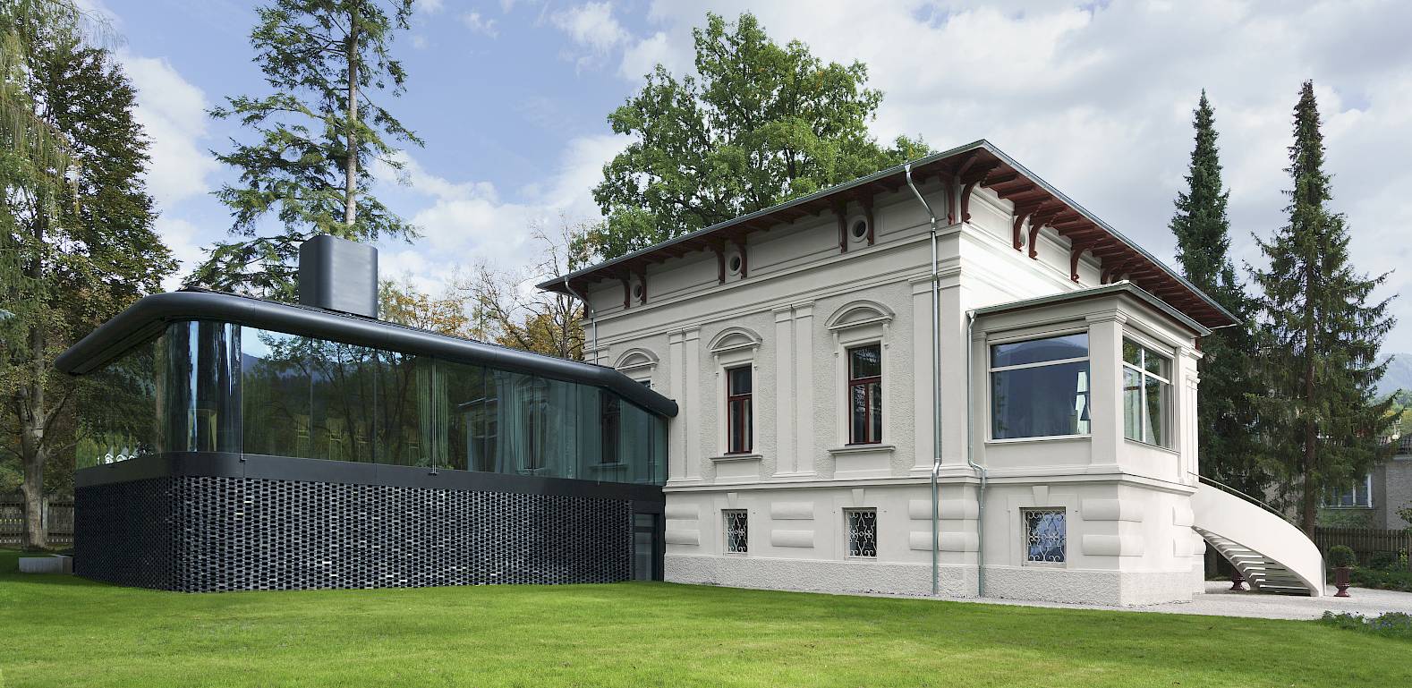 Historische Villa mit modernem Anbau, eingebettet in eine grüne Landschaft unter einem bewölkten Himmel.