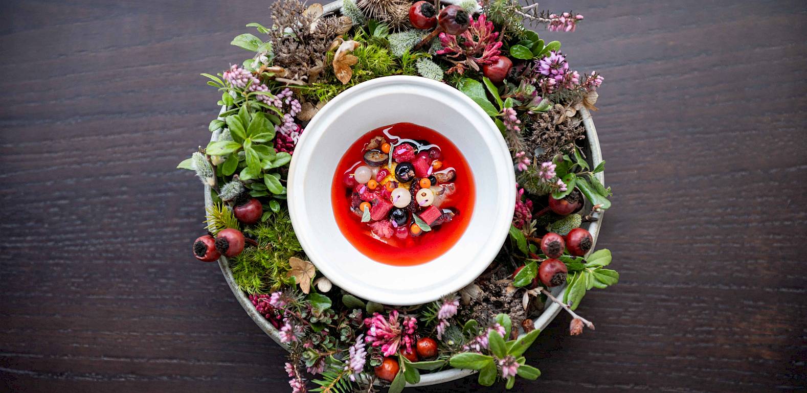 Eine Schüssel roten Fruchtpuddings, dekoriert mit frischen Beeren und grünen Blättern auf einem Holztisch.