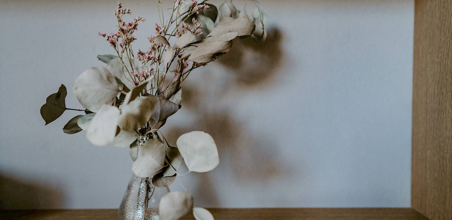 Vaso con fiori secchi e foglie su uno scaffale di legno davanti a una parete neutra.