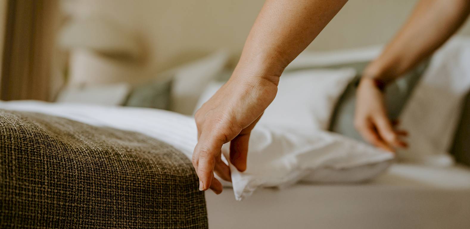 Eine Hand, die ein Bettlaken glättet, während ein Bett gemacht wird. Der Fokus liegt auf der Präzision der Bewegung.