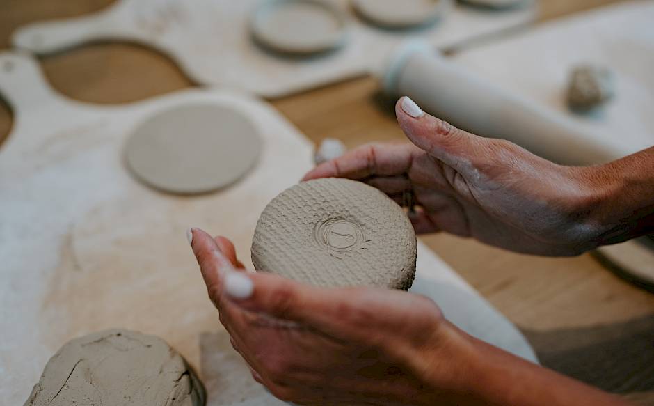 Hands holding a round clay disc, surrounded by other clay pieces on a table.