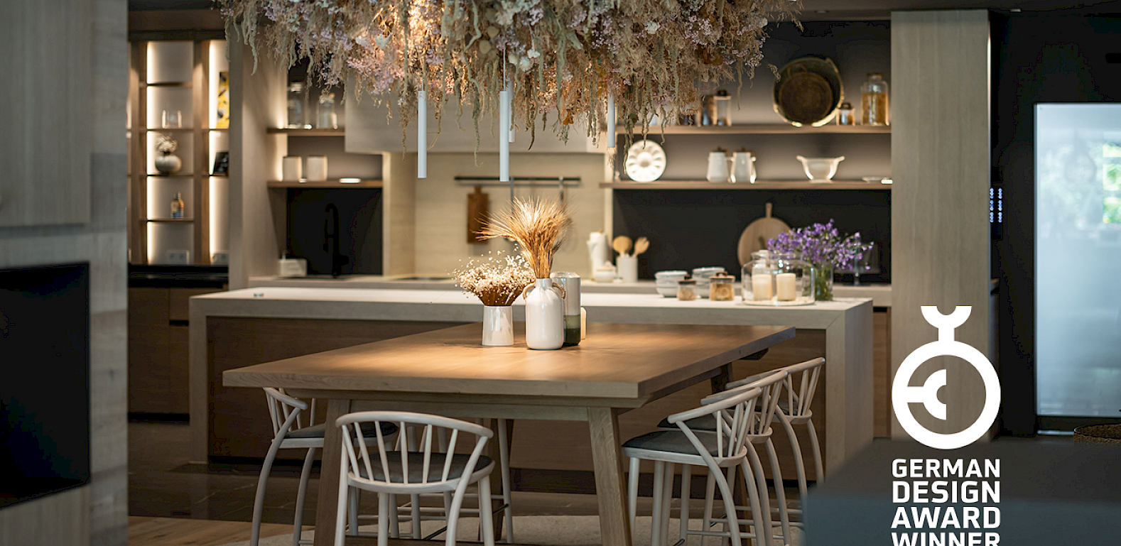 Moderne Küche mit großem Holztisch, dekorativen Trockenblumen und "German Design Award Winner 2025"-Logo.