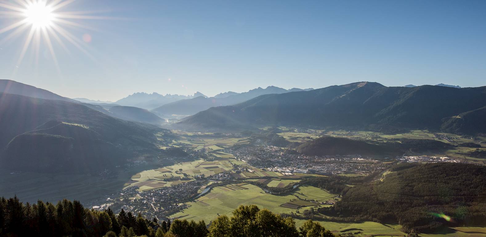 Panoramablick auf ein Tal mit Wäldern und Bergen unter einem klaren Himmel und strahlender Sonne.