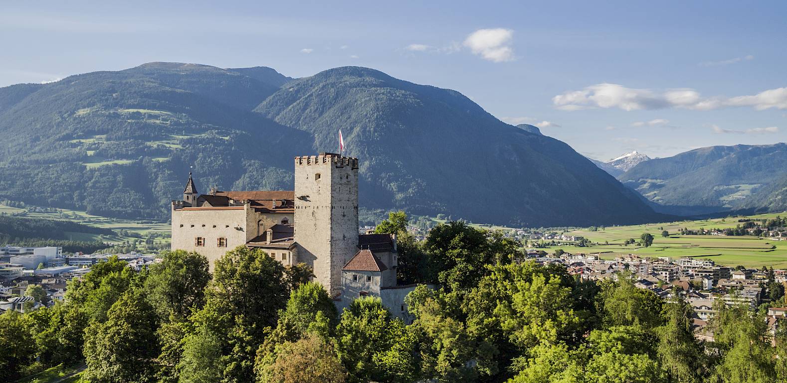 Schloss auf einem bewaldeten Hügel, umgeben von Bergen und Wiesen unter blauem Himmel.