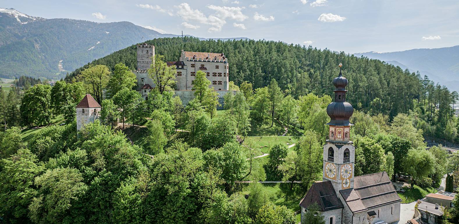 Grünes Hügelpanorama mit Kirche und mittelalterlichem Schloss, umgeben von Wald und Bergen im Hintergrund.