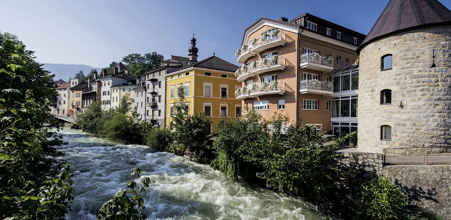 Fluss fließt an bunten Gebäuden und einem runden Turm in einer malerischen Stadtlandschaft vorbei.