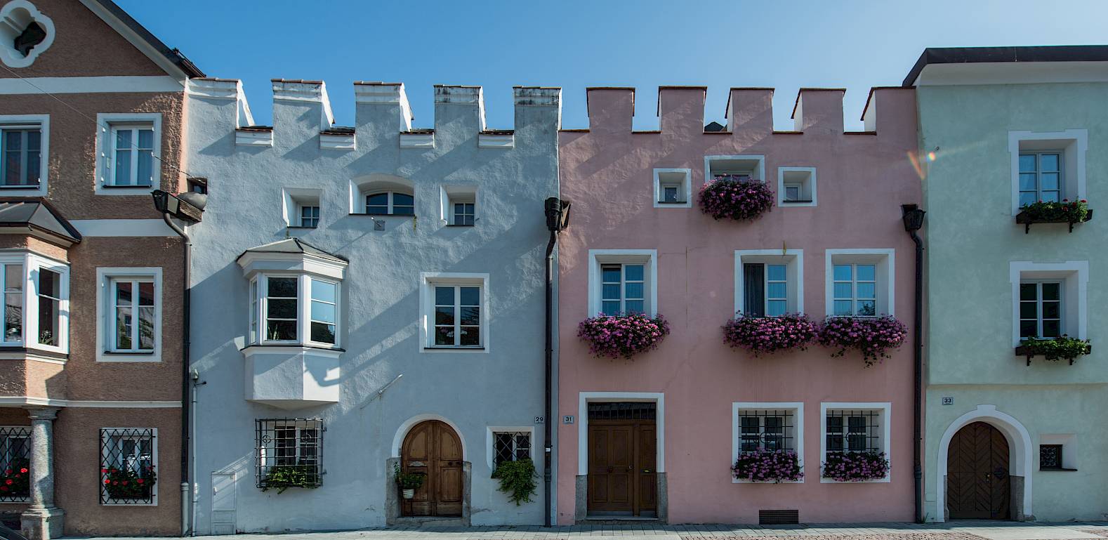 Pastellfarbene Gebäude mit Zinnen und Blumenkästen an Fenstern in einer sonnigen Kopfsteinpflasterstraße.