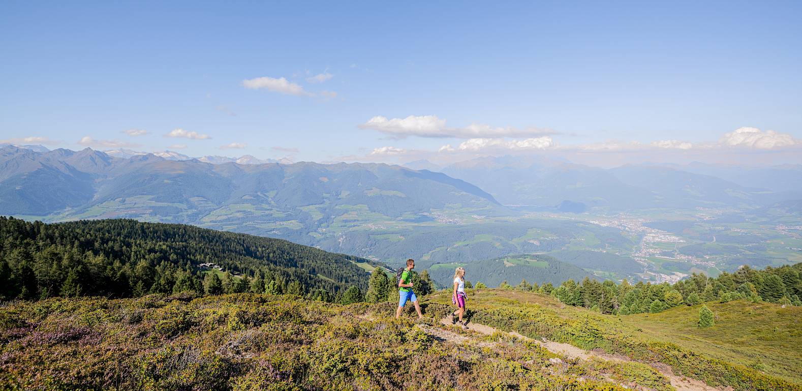 Zwei Wanderer auf einem Bergpfad mit weitem Panoramablick auf das Tal und entfernte Bergketten.