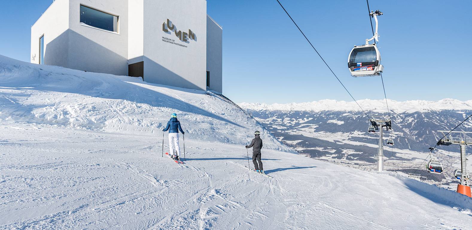 Zwei Skifahrer auf einer verschneiten Piste, daneben ein modernes Gebäude und ein Skilift unter blauem Himmel.