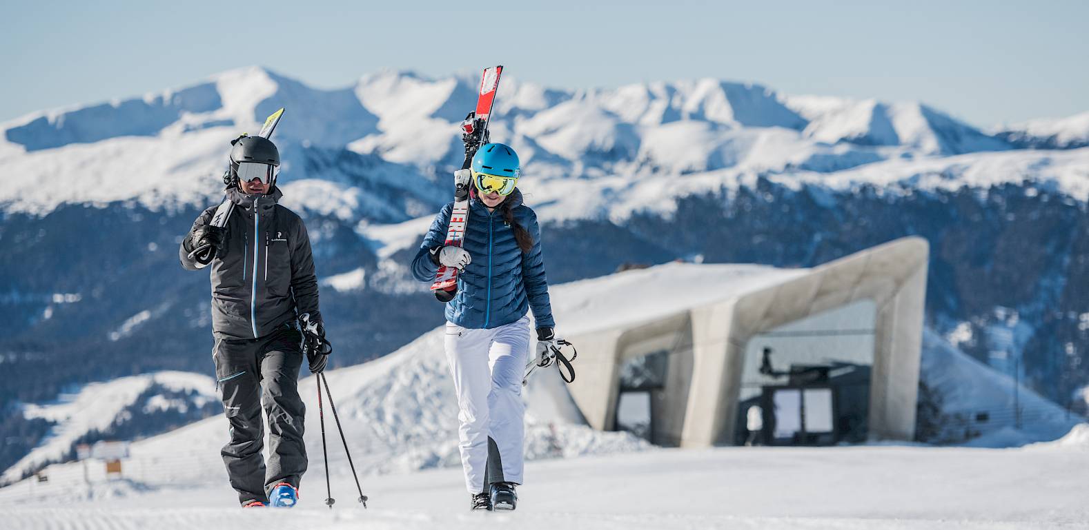 Zwei Personen mit Skiern spazieren auf einem verschneiten Berg vor einem modernen Gebäude in den Alpen.