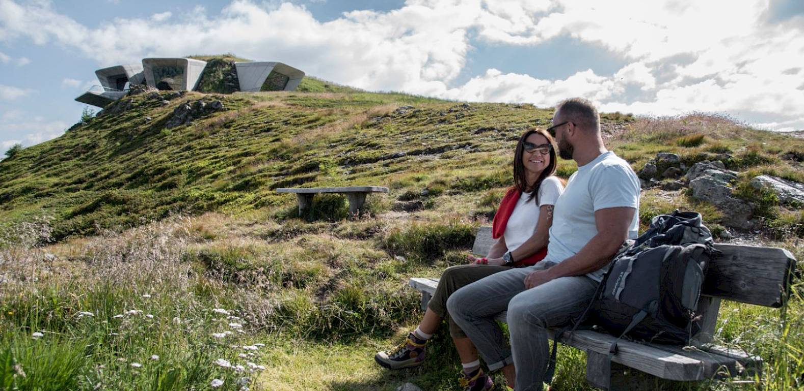 Paar sitzt auf einer Bank in einer grünen Berglandschaft, im Hintergrund moderne Architektur unter bewölktem Himmel.
